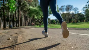 Mladá žena v sportovním oblečení s úsměvem běží po zpevněné cestě v zeleném parku. Její odhodlání odráží cíle průvodce pro začátečníky v běhu.