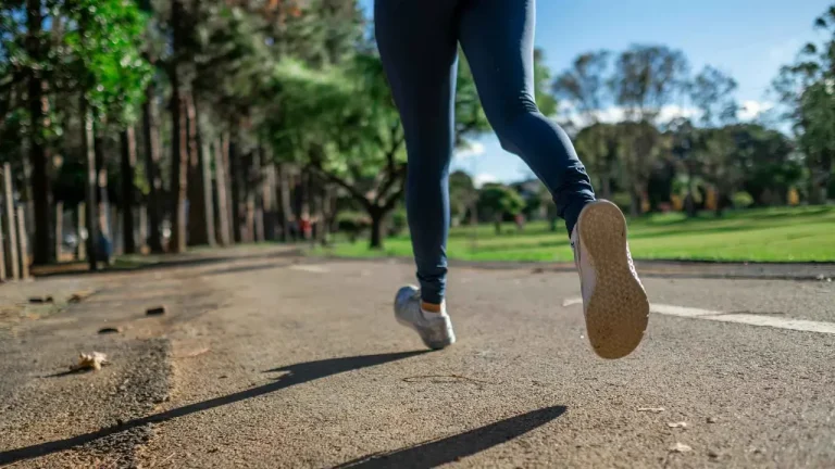 Mladá žena v sportovním oblečení s úsměvem běží po zpevněné cestě v zeleném parku. Její odhodlání odráží cíle průvodce pro začátečníky v běhu.