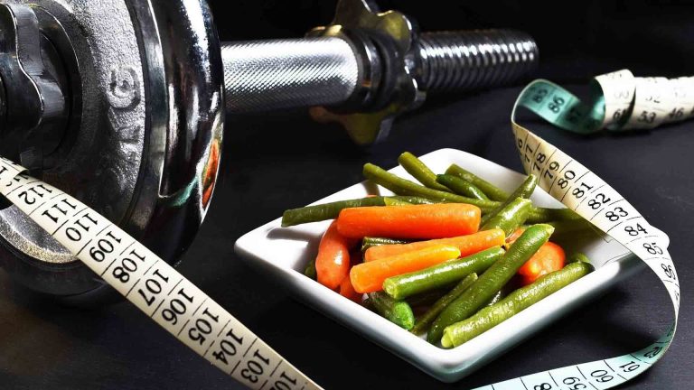 eine Schüssel mit frischem, nahrhaftem Essen neben einer Hantel und einem Maßband.