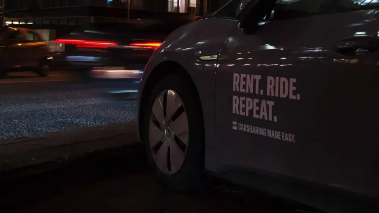 Un uomo sorridente consulta un'app di noleggio auto sul suo smartphone, con diverse opzioni di car sharing disponibili in Italia sullo sfondo urbano.