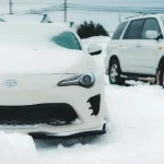 Auto sa opatrne pohybuje po zasneženej ceste s rozsvietenými svetlami, čo znázorňuje dôležité tipy na bezpečnú jazdu v zime.