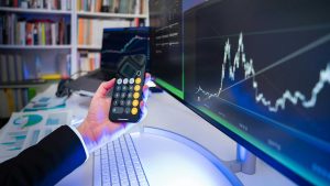 Osoba obserwująca wykres giełdowy, trzymająca telefon z otwartą aplikacją kalkulatora.